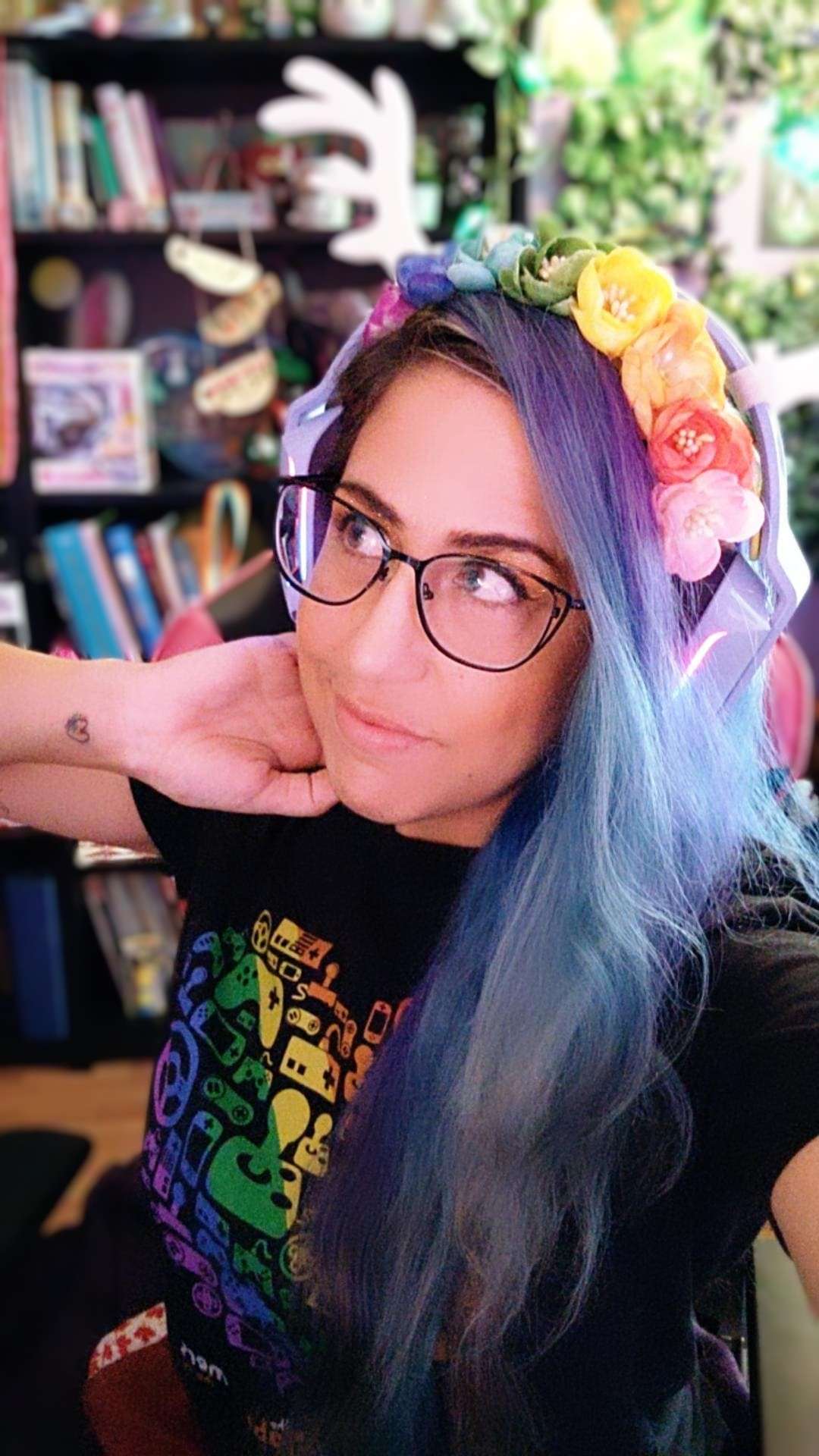 Person wearing glasses, wearing a shirt that has multiple video-game type controllers on it, wearing a rainbow flower crown