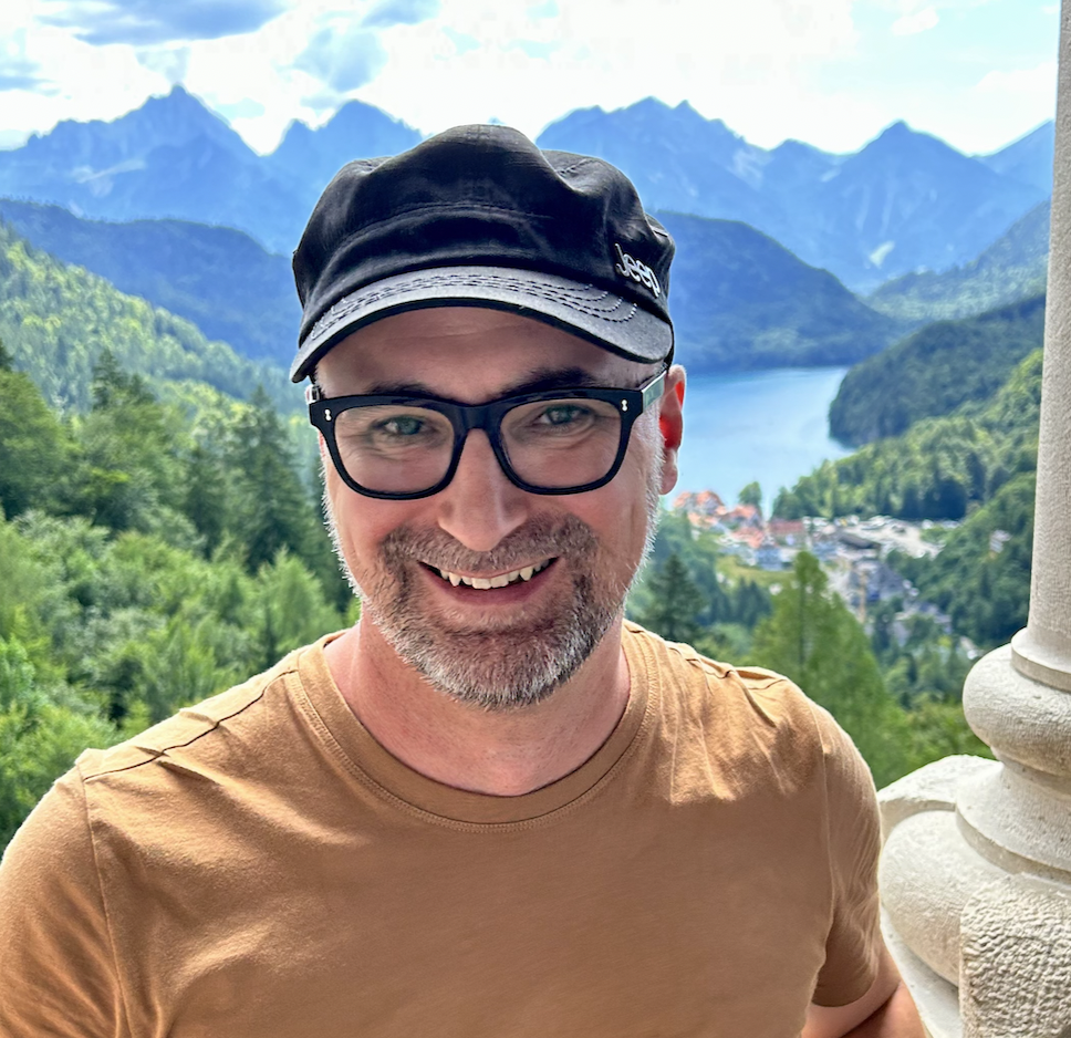 Derek Gour, clad in stylish glasses and a baseball cap, smiles into the camera, distant mountains, and an alpine village on a lake in the distance.