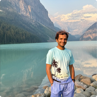 Jigar Chandra standing on a rocky beach in front of a clear blue lake and mountains