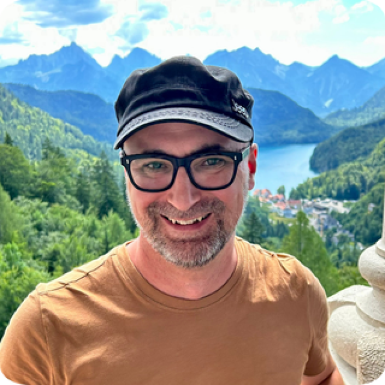 Derek Gour standing in front of a forested lake with mountains in the background. 